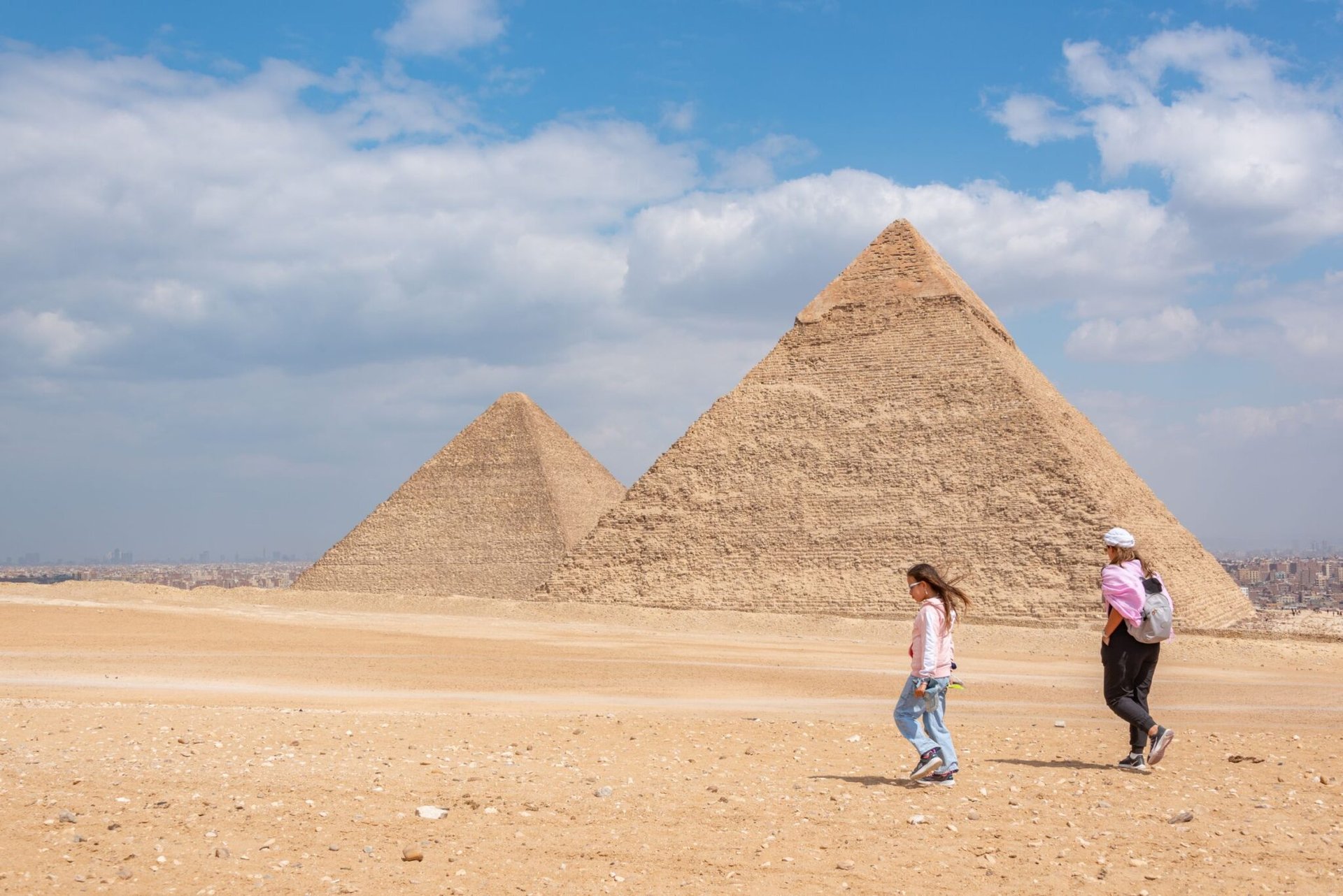 Two women walking near the iconic pyramids of Giza, Egypt, under a bright sky.