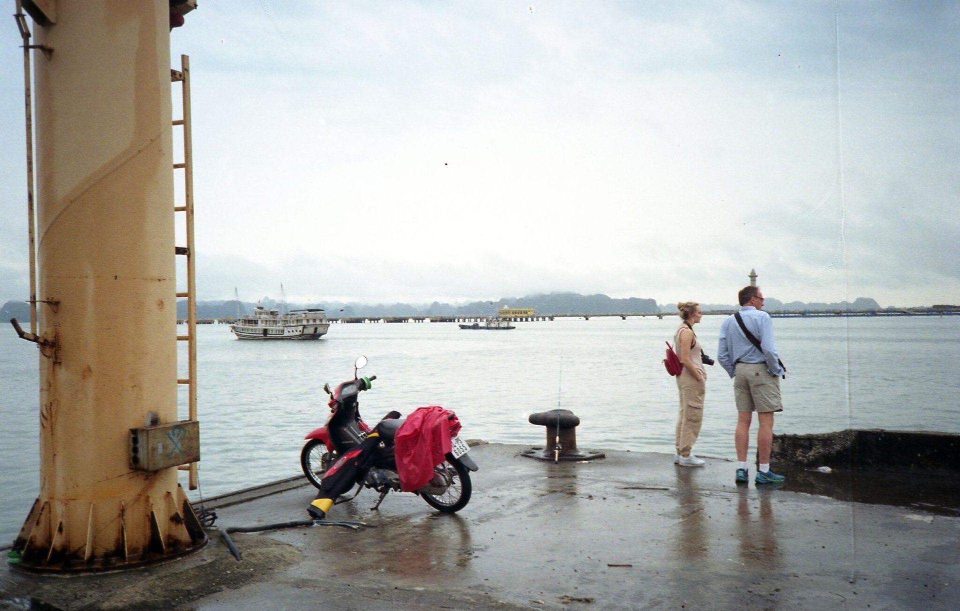 Couple stands on a pier at Ha Long Bay, Vietnam, with a ship in the distance.