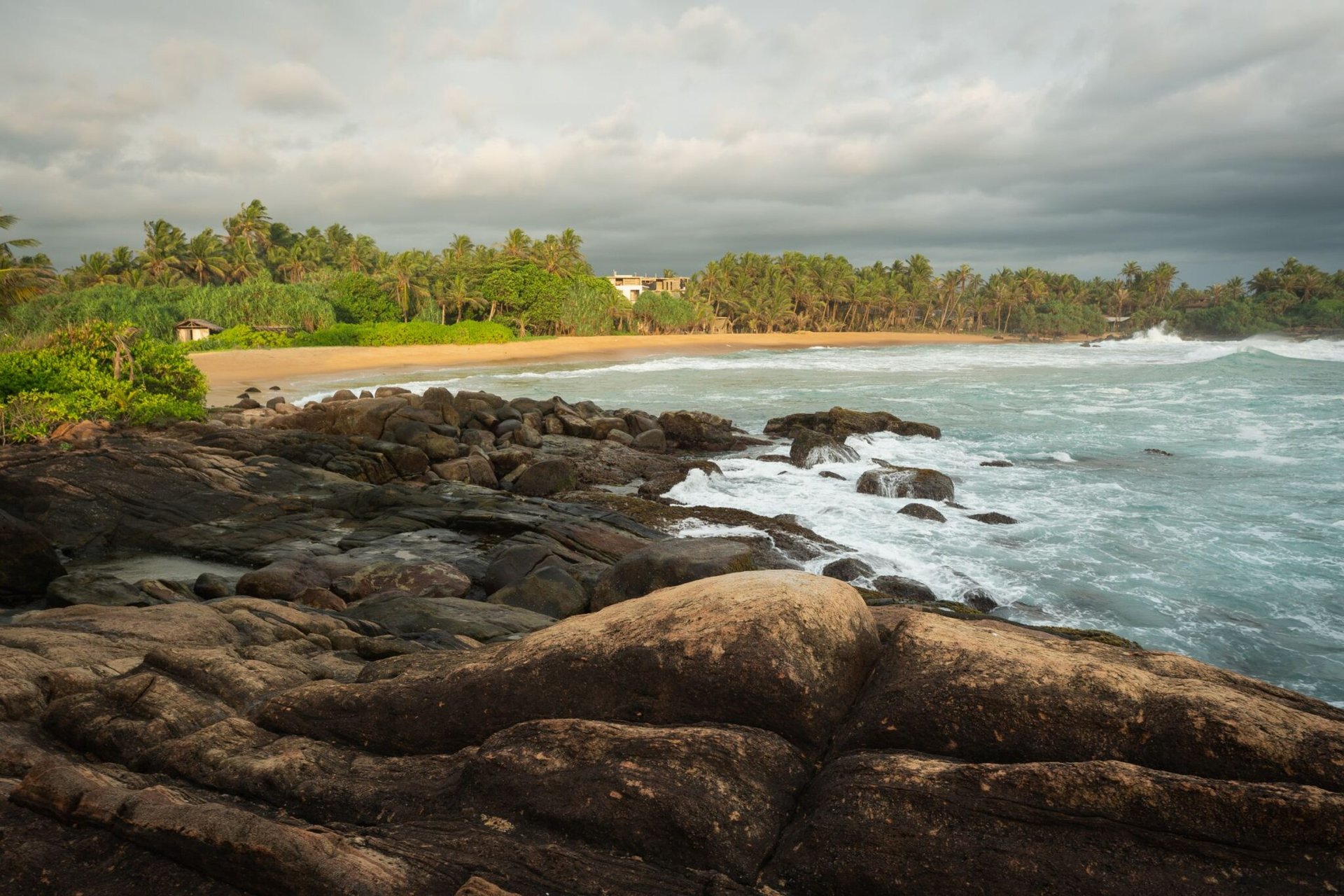 Explore a serene tropical beach in Sri Lanka with rocky shores and lush greenery under a dramatic sky.