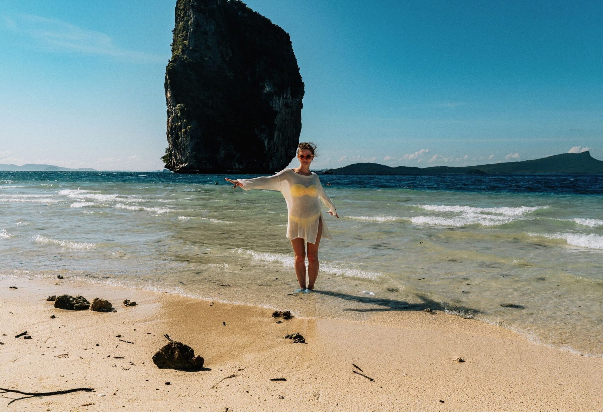 A woman stands on a sunny beach in Phang Nga, Thailand, with a towering rock formation in the background.