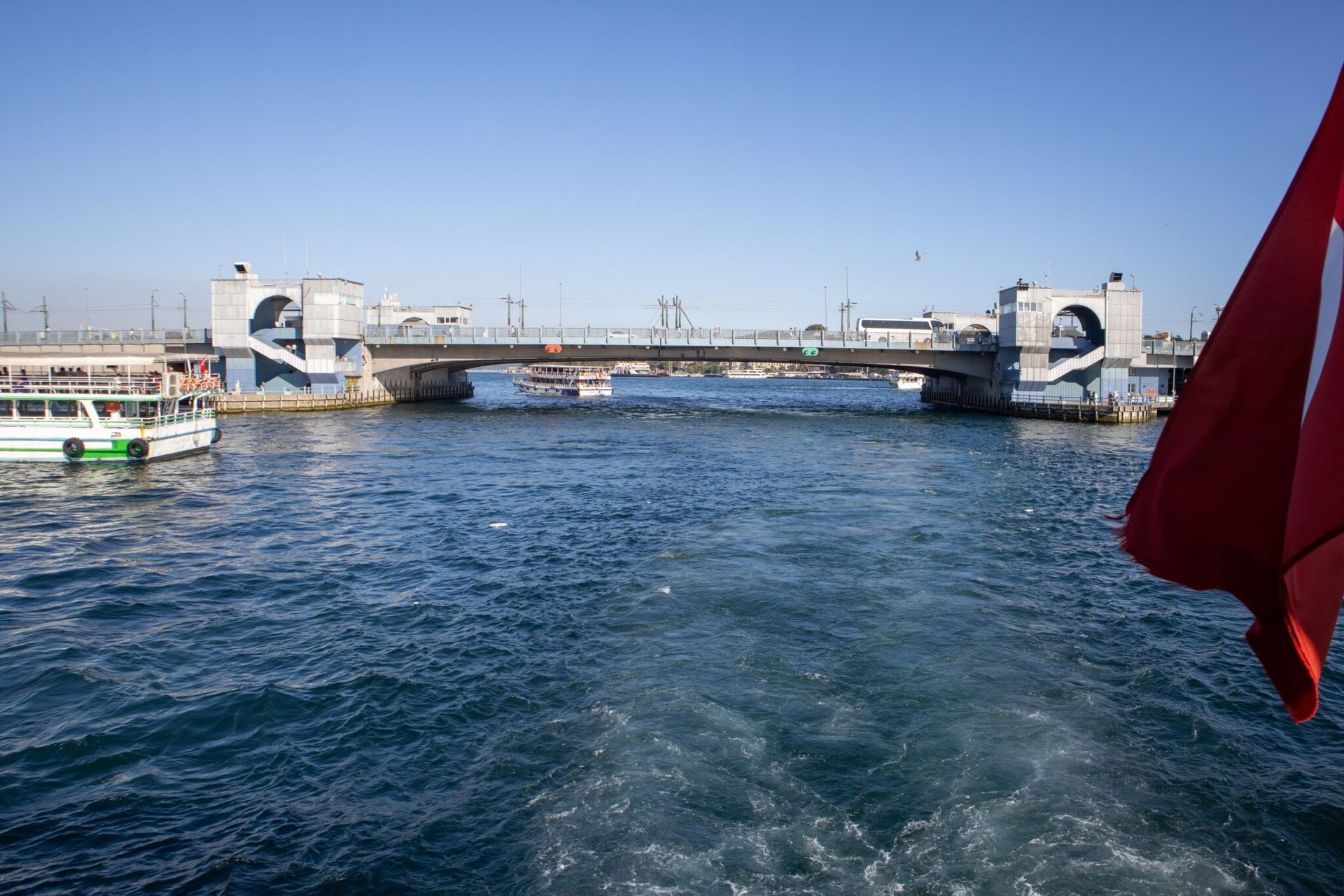 Sunny day view of Galata Bridge spanning the Bosphorus in Istanbul, capturing boats and a Turkish flag.