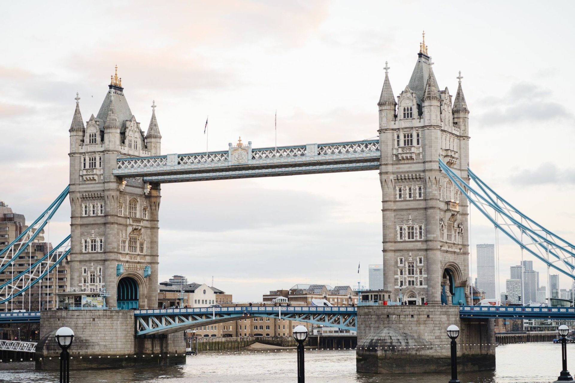 Historic suspension bridge with towers connected by walkways located over calm river against residential buildings in London city against cloudy sky